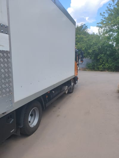 A side view of a white box truck is captured in this image. The truck is parked on an asphalt surface, possibly a street or parking lot, with a wooded area and a chain-link fence visible in the background. The sky above is partly cloudy. The truck features a large white cargo box and an orange cab. A tire and part of the truck's chassis are visible on the lower left side. The scene appears to be outdoors during the daytime, with natural lighting. The location is identified as Alchevsk, Ukraine.