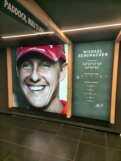 The image captures an indoor display at the Red Bull Ring complex in Spielberg, near Flatschach, Austria. On the left, a large, brightly illuminated portrait depicts Michael Schumacher, smiling and wearing a red racing cap. To the right, an illuminated dark green panel displays white text including "MICHAEL SCHUMACHER," "#1 WELTMEISTER" with seven laurel wreath icons, "STATISTIK UND REKORDE," and "REKORDE IM SPIELBERG," concluding with a white signature. Both panels are framed by vertical wooden beams. An overhead sign, partially visible, reads "PADDOCK, BULL'S LANE," suspended from a wooden ceiling beam. Linear LED light strips illuminate the displays and the dark, textured ceiling. The floor is covered with dark tiled squares featuring light-colored grout lines. No individuals are present.