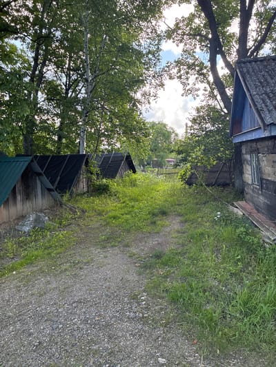 A dirt path leads through a grassy area with several small, rustic outbuildings on the left and a dilapidated wooden structure on the right. Tall trees with green foliage frame the scene, and the sky is partly cloudy. The outbuildings appear to be sheds or small shelters, with one having a green metal roof and others covered in black material. The wooden structure on the right is weathered, with visible planks and a single window. A wooden fence is visible in the background, and a distant industrial chimney can be seen through the trees. The environment suggests a rural or semi-abandoned setting.