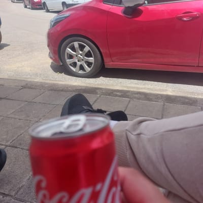 In Ablanitsa, Bulgaria, a person is seated on a sidewalk, holding a Coca-Cola can. The can is red with white lettering and is positioned in the foreground, partially obscuring the person's legs which are clad in beige pants and black athletic shoes. Behind the person, a red car is parked on the street, its front wheel and side visible. The sidewalk is paved with grey square tiles. The background shows a paved street and other vehicles.