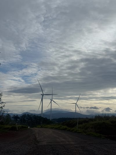 The image shows a landscape in Phát Chi, Vietnam, featuring wind turbines against a cloudy sky. A rough, dark road runs along the right edge of the frame, with vegetation and trees bordering it. In the distance, layers of mountains are visible through gaps in the clouds. Three wind turbines stand prominently in the middle of the image, their blades extending into the sky. The sky is predominantly gray with textured clouds, with hints of blue peeking through.