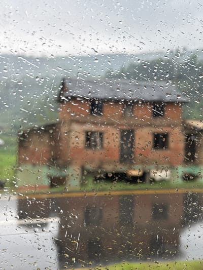 The image shows a blurred view through a rain-streaked glass surface. In the background are buildings, indistinct but appearing to be in an urban setting, with one building showing a grid-like pattern of dark window openings on a lighter brick facade. A vertical yellow stripe-like feature is also visible. Given the prompt's context, the location could be Tharpu, Nepal. The raindrops on the glass obscure the details of the city beyond, creating a distorted and impressionistic view. No people are visible.