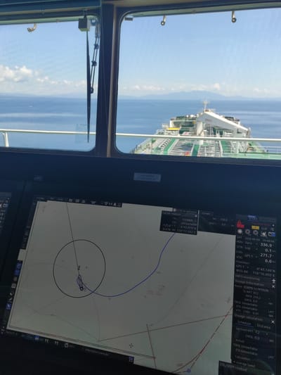 This vertically oriented image captures a view from the bridge of a vessel, looking out to sea on the left and at an Electronic Chart Display and Information System (ECDIS) on the right.

Through the ship's window on the left, the clear blue sky and calm blue ocean are visible, with distant land or mountains faintly outlined on the horizon. The foredeck of a large vessel, likely a tanker, with its intricate piping and structures, extends into the frame, suggesting the ship is at sea in bright daylight conditions. Window frames and a windshield wiper are also visible.

The right side of the image is dominated by the ECDIS screen, displaying navigation and route information. The brand "JRC" is visible on the display. Key data points indicate the date as 2026-06-04, time as 10:44:04 LMT, and the vessel's position as 4°43.149' N, 118°16.955' E, which places it near Kunak, Malaysia. The Speed Over Ground (SOG) is 0.0 kn, and Speed Through Water (STW) is 0.1 kn, suggesting the vessel is stationary, possibly at anchor or drifting slowly.

The screen also shows the ship's heading (HDG Gyro) at 336.9° and Course Over Ground (COG GPS 1) at 271.7°. Route information indicates a journey from "IDBPN to MYKUN" with a destination waypoint (WPT 012). "TWOL Unreachable" and "ETA Unreachable" are displayed, consistent with a stationary vessel. The chart view on the ECDIS shows the vessel's current position, a planned blue route line, and a circular safety zone around the ship, with other targets or vessels indicated.

Other visible text on the screen includes "PRIMARY ECDIS," "Current Set 156.9 kn," "DRIFT 0.0 kn," "DISP brightness," "Cursor readout," "TTG: Unreach," "125.6° T," "0.478 NM," "Vector(T) 6 min," "Filter AIS GND TT," "ANT1," and "Information overscale." The map scale is "1:6,000." The clear weather and detailed navigational display suggest routine maritime operations.