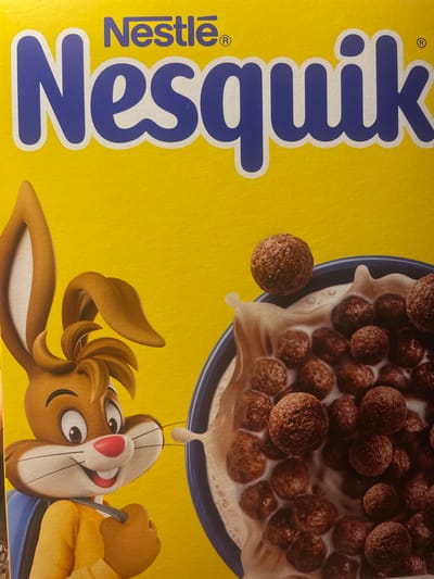 A bright yellow Nesquik cereal box features the brand name "Nesquik" in large, blue letters. A bowl of chocolate cereal in milk is shown at the top right, with one cereal ball hovering above. Below, the Nesquik bunny character is depicted upside down, appearing to catch a drop of milk. The overall image is brightly lit and focused.