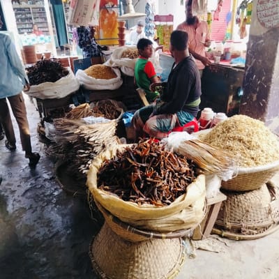 The image depicts a scene at a market in an unidentified city in Bangladesh. In the foreground, there are several woven baskets and sacks filled with dried goods, including what appears to be dried fish or seafood. The largest basket contains a pile of dark brown, elongated items. Another basket is filled with lighter-colored, smaller items, likely also dried fish. There are bundles of dried plant stems or reeds among the baskets.
In the background, people are engaged in commerce. Two men are sitting and talking near the dried goods. A man in a pink shirt appears to be serving or interacting with another person at a stall. Further back, a building or store is visible with shelves stocked with various items. The environment is bustling with activity, typical of a market scene. The floor is wet and worn. A writing on a board is visible.