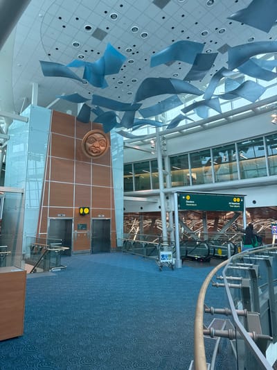 The image depicts the interior of a modern airport terminal in Richmond, Canada. The scene is characterized by a spacious, well-lit environment with a high ceiling. Suspended from the ceiling are numerous abstract, blue metallic sculptures, creating a dynamic visual display.

The main focus of the image is a wall featuring two elevators. Above the elevators, a large, circular wooden sculpture resembling a face with tribal or indigenous features is mounted. To the left of the elevators, there are two illuminated yellow buttons, one with a person icon and an upward arrow, and another with a person icon and a downward arrow, likely indicating elevator controls.

A prominent green directional sign is visible, indicating "All Departures" with an upward arrow and "Elevators" with a leftward arrow, both in English and French. A person in a green jacket is standing near the escalators, which are also visible in the background.

The floor is covered with a blue carpet featuring a circular patterned design. Glass panels and metal railings line the walkways and staircases, adding to the modern aesthetic of the terminal. The overall impression is one of efficiency and contemporary design. The lighting suggests it is daytime.