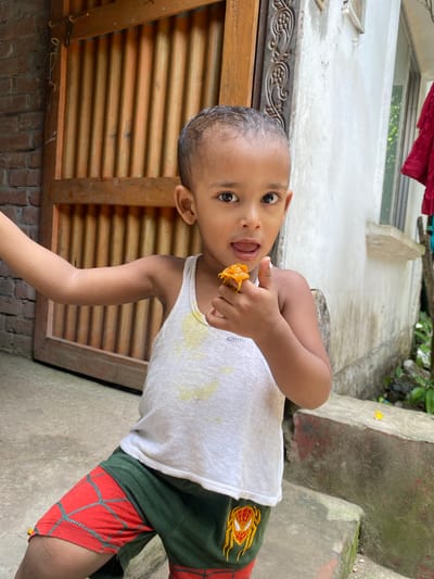 A young child stands outside near a wooden door and a concrete structure. The child has dark hair and skin, wearing a white tank top and dark green shorts with red, patterned underpants showing. The child is holding a piece of orange food to their mouth, looking at the camera. The location is potentially Hizla, Bangladesh.