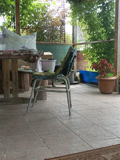 The image shows a covered outdoor porch with a tiled floor and wooden support beams. A wooden table covered with a patterned tablecloth is to the left, with a white pillow with floral print on it. A metal chair with a patterned cushion is in the center of the frame. To the right, a red plant in a brown pot sits on the tiled floor. In the background, there is greenery and some blue material. The scene appears to be in a rural or suburban setting.