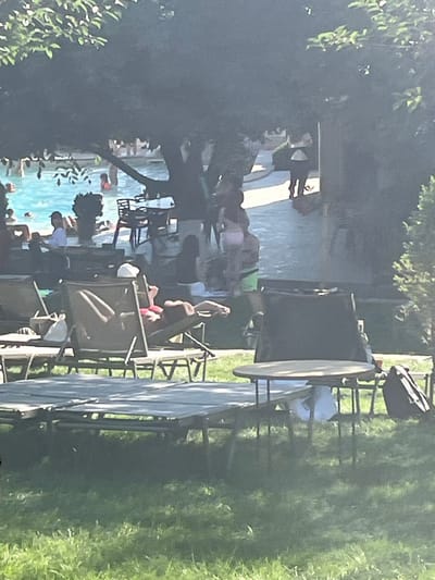 The image depicts an outdoor scene with a swimming pool and people relaxing on lounge chairs under the shade of trees. The environment suggests a recreational area, possibly a resort or a park. In the foreground, several empty lounge chairs are visible on a grassy area, with one chair occupied by a person. The swimming pool is in the background, with several individuals swimming or wading. Some people are also seen walking on a paved path near the pool. The overall atmosphere appears to be a sunny day at a leisure facility. The location is not specified as Stăuceni, Moldova.