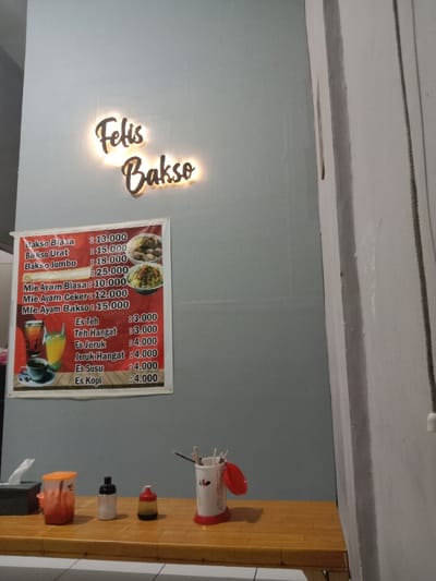 The image shows a restaurant in Kecamatan Brangsong, Indonesia, located at latitude: -6.944, longitude: 110.221. The restaurant is named "Felis Bakso" and features a bright red and white signboard with a price list for bakso (meatball soup), mie ayam (chicken noodle soup), and various drinks. The signboard is mounted on a pale grey wall. To the right of the signboard is a small table with a red plastic container of chopsticks, a white bottle with a red cap, a red bottle with a white cap, and a red container with a white lid. The table is wooden and has a light brown finish. There is a white wall to the right of the table. The environment is clean and well-lit.