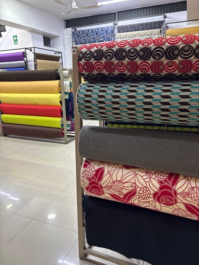 The image shows a retail store with rolls of fabric stacked on display shelves. The fabrics feature various patterns and colors, including geometric designs, floral prints, and solid hues. Some of the rolls are brightly colored, while others are more muted. The store appears to be well-lit, with fluorescent lights on the ceiling and natural light coming from a window or doorway in the background. There are no people visible in the image. The location is identified as La Victoria, Peru.