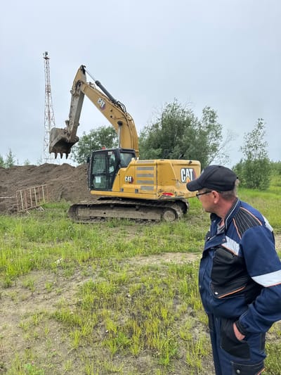 In Usinsk, Russia, a man wearing a dark blue work jacket with orange and black accents and a black baseball cap stands with his hands in his pockets, facing away from the camera to the right. He is in a grassy area with patches of dirt. Behind him is a large, yellow Caterpillar excavator, with its bucket raised. The excavator is positioned next to a pile of dark soil. In the background, a tall, lattice tower with a small antenna at the top is visible, along with several trees and an overcast sky.