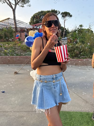 A woman with brown hair and sunglasses is standing outside, holding a popcorn container and eating popcorn. She is wearing a black crop top and a denim skirt. There are balloons and bubbles in the background. The location appears to be a park or outdoor venue, possibly in Sant Vicenç de Montalt, Spain, given the presence of lush vegetation and what seems to be a modern structure in the background, consistent with a recreational area. The sky is clear with a soft light, suggesting it might be late afternoon or early evening.