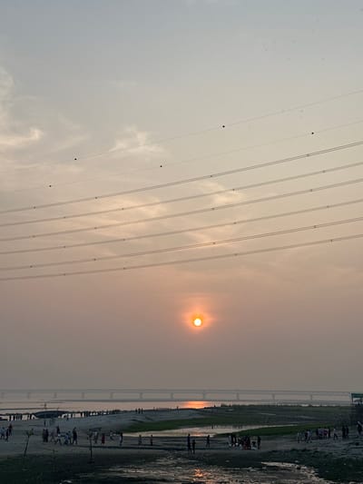 The image captures a wide outdoor scene at sunset in Kumarvog, Bangladesh. A prominent orange-yellow sun is positioned low on the horizon, slightly right of center, casting a bright reflection across a wide body of water in the middle ground. A long, dark truss bridge spans this water body in the distant background. The sky above is hazy, transitioning from a light orange near the horizon to a pale grey-blue towards the top. Multiple parallel overhead power lines, each fitted with several small black spherical markers, traverse the upper half of the frame. The foreground and midground consist of sandy and muddy riverbanks or char lands, interspersed with shallow water channels and some patches of green vegetation. Numerous individuals and small groups are dispersed across these land areas and shallow waters, engaged in walking and congregating. A dark, hull-shaped boat is beached on the left-hand bank. The scene is illuminated by the late afternoon or dusk light.