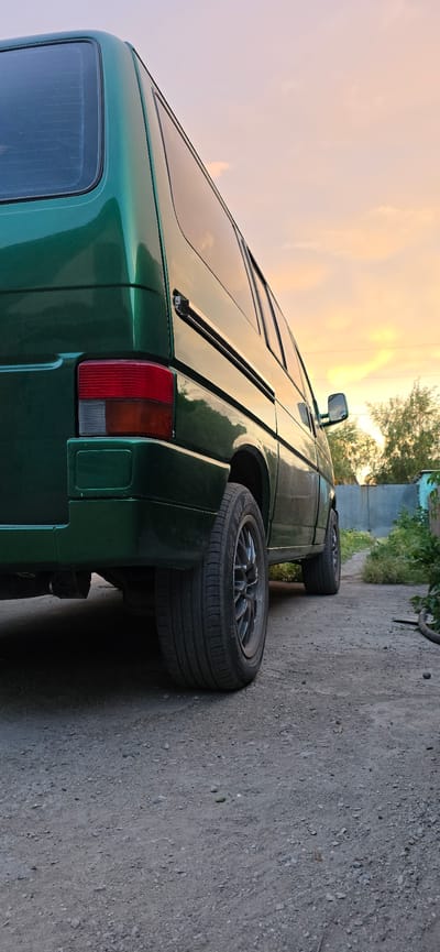 This media file shows a green van parked on a dirt road. The van is seen from the rear, with its taillight and a portion of its side visible. The van has alloy wheels and the tires are clearly visible. In the background, there are some trees and a building, suggesting an outdoor setting. The sky is visible, with some clouds, and the light suggests it is either sunrise or sunset. There is no visible text in the image. The setting is outdoors on a dirt road. The van is the main object. There are no people visible. The overall mood of the image is peaceful and serene, likely due to the soft lighting. The location context provided, Saran city administration, Kazakhstan, is not directly inferable from the image itself, but provides additional information about the scene.