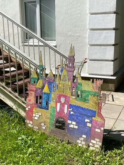 An outdoor scene features a colorful, cutout wooden castle placed in a grassy area next to an external staircase. The castle is painted with various bright colors, including red, purple, blue, green, and yellow, depicting towers, windows, and brickwork. A shopping cart is visible behind the castle, with its handle extended. The staircase, made of metal with some rust and carpeted treads, leads upwards along a textured white building wall. A window with blinds is present on the building. The scene is bathed in sunlight, casting shadows on the ground. The location is identified as Obninsk, Russia. There are no people visible in the image.