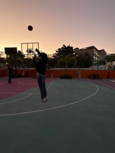 A person in a black jacket and jeans is throwing a basketball on an outdoor court at sunset. The court is painted with green, pink, and white lines. In the background, there are trees, buildings, and a fence with writing on it. Another person is standing near the edge of the court. The sky is a gradient of orange and purple hues.