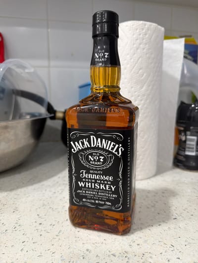 A close-up, angled shot shows a bottle of Jack Daniel's Tennessee Sour Mash Whiskey lying horizontally on a white speckled countertop. A roll of white paper towels is behind the bottle, along with a dark-colored can. A partial view of a silver-colored bowl is visible in the lower foreground. The lighting is bright and the environment appears to be a kitchen or similar food preparation area. The location is not Pickering, Canada.