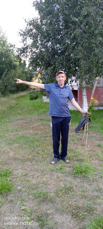A middle-aged man wearing a blue patterned polo shirt, dark sweatpants, a light-colored baseball cap, and sandals stands on a grassy area. He is pointing his left arm to the left, with his palm open. In his right hand, he holds a black plastic bag, a thin wooden stick, and a small yellow flowering plant. Behind him, a birch tree with a white trunk stands to the right, with a building visible through its leaves. The ground is covered in dry grass and weeds. The image appears to be taken outdoors during the day, with natural light. The timestamp in the lower left corner indicates the date and time as 2026/07/29 18:04, and the device as Infinix NOTE 30i.