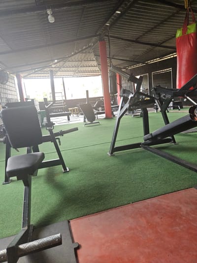 The image shows the interior of a gym located in Perímetro Urbano Cúcuta, Colombia. The scene is dominated by various pieces of weightlifting equipment. Black metal gym equipment is placed on green artificial turf. A red punching bag hangs from the ceiling in the upper left corner. The building has a corrugated metal roof. Natural light enters from windows along the back wall. The overall impression is of a functional, well-equipped fitness facility.