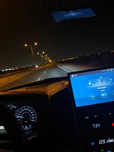 The media file captures a view from inside a car at night, likely on a highway in Umm Salal Ali, Qatar. The scene depicts a vehicle moving or parked on a well-lit road, with numerous streetlights visible in the distance creating a bokeh effect.

Inside the car, an infotainment screen is prominently displayed, showing music playback. The song title "Мальборо" (Marlboro) by "SAYAN" is visible, along with an equalizer graphic and media control icons. The time on the screen reads 19:35, and "BT" indicates Bluetooth connectivity. Adjacent to the infotainment screen, another display shows "734" and a climate control setting of "22.0" degrees Celsius with a fan icon.

The car's instrument cluster, specifically the speedometer, is partially visible at the bottom right. The needle rests at 0 km/h, suggesting the vehicle is stationary or moving very slowly at the moment the photo was taken. The speedometer scale goes up to 240 km/h, and a fuel gauge and a voltmeter showing "19V" are also present. A reflection of the infotainment screen, with distorted text like "Wourgobo," is visible on the windshield. The overall atmosphere suggests a calm night drive.