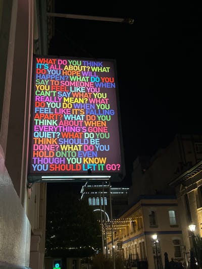 This nighttime image captures an illuminated art installation in Fan Tan Alley, Victoria, British Columbia, Canada. A large, rectangular sign, oriented vertically and mounted perpendicularly to a light-colored building on the left, displays a series of introspective questions in bright, multi-colored capital letters (red, orange, yellow, green, blue, purple, pink) against a dark background. The text reads: "WHAT DO YOU THINK IT'S ALL ABOUT? WHAT DO YOU HOPE WILL HAPPEN? WHAT DO YOU SAY TO SOMEONE WHEN YOU FEEL LIKE YOU CAN'T SAY WHAT YOU REALLY MEAN? WHAT DO YOU DO WHEN YOU FEEL LIKE IT'S FALLING APART? WHAT DO YOU THINK ABOUT WHEN EVERYTHING'S GONE QUIET? WHAT DO YOU THINK SHOULD BE DONE? WHAT DO YOU HOLD ONTO EVEN THOUGH YOU KNOW YOU SHOULD LET IT GO?" To the right, a street or alley recedes into the distance, lined with buildings featuring architectural details and illuminated by overhead string lights and several street lamps. A distant, taller building with internal lighting is visible in the background, along with a tree. Indistinct human figures are faintly discernible on the right side of the street near the bottom of the frame.