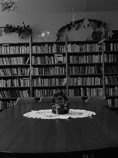 A black and white photograph shows an interior scene with a large wooden table in the foreground, covered with a white tablecloth and centered with a bouquet of flowers in a basket. The table is set with several chairs, positioned against a long, wooden bookcase filled with books. The bookcase is against a wall, decorated with a vine of leaves. The scene is likely located in a library or meeting room. The location, based on coordinates, is Berehove, Ukraine.