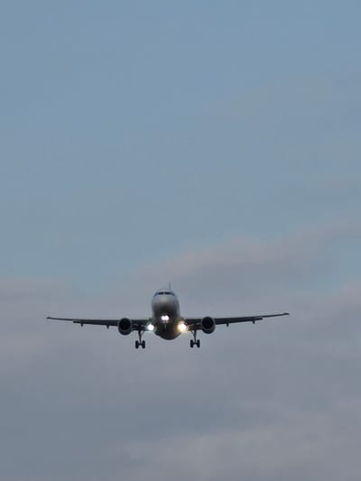 An airplane is captured in flight against a cloudy sky, its landing gear extended, indicating it is likely on approach for landing. The aircraft is silver and appears to be a commercial passenger jet. The lights on the front of the plane are illuminated. The image was taken in the vicinity of Kelsterbach, Germany, which is near Frankfurt Airport (FRA), a major international airport. The sky is a muted blue with some diffused clouds.