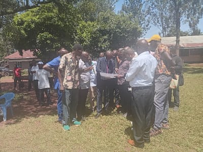 The scene, located in Idakho South ward, Kenya, depicts an outdoor gathering of approximately 15 to 20 adult males on a grassy area under clear daylight conditions. In the central foreground, a cluster of ten men is intensely focused on several white documents or maps held by individuals within the group, including one man in a dark suit, white shirt, and patterned tie. Another man, wearing a light blue collared shirt and dark trousers, stands with his back to the viewer, also examining the documents. A man in a floral-patterned short-sleeved shirt and dark trousers is positioned to the left of the central group, looking downwards, while another man in a yellow cap and patterned shirt is on the right.
To the left of the main cluster, several men stand observing, including individuals in a blue shirt and white t-shirts. A single light blue plastic chair is positioned on the grass in the lower-left corner. A black sedan is partially visible behind the peripheral group of men, adjacent to a building with a red-tiled roof. Dense green foliage from large trees forms the dominant background element across the upper frame. In the far-right background, a single-story building with light-colored walls and a darker, possibly corrugated, roof is visible. The ground throughout the visible area is covered in short, green grass.