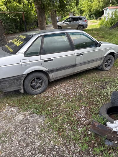 In the city of Ayet, Kazakhstan, a silver sedan is parked on a gravel and grass lot. The car has a yellow sticker on the rear window that reads "YUNHA klub.kst Kostanay". A second, darker colored SUV is parked behind it. The area is covered in green trees and grass. In the foreground, there are several discarded tires and a rusty piece of metal.