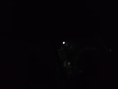 This is a dark, out-of-focus image taken at night, likely in or around the city of Ақсай, Kazakhstan. The scene is dominated by blackness, with a few faint, blurred lights visible. One prominent, slightly larger light source is located near the center-right of the image, appearing somewhat diffused. Several other smaller, indistinct white or yellowish orbs are scattered in the lower right portion of the frame. The overall impression is one of very low light conditions, possibly indicating an outdoor environment at a considerable distance from significant artificial illumination, or a heavily obscured light source. The blurry nature of the image prevents any identification of specific objects, people, or activities.