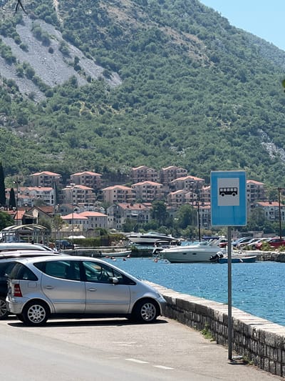 The image depicts a scenic view of Risan, Montenegro, with a steep, verdant hillside covered in trees and rocks dominating the background. A cluster of buildings with terracotta roofs is nestled at the base of the hill, facing a bright blue body of water, likely the Bay of Kotor. Along the waterfront, a paved walkway runs parallel to the water's edge, with a low stone wall separating it from the sea. Several boats and yachts are moored in the water.

On the walkway, a silver car is parked, its side visible. A bus stop sign, featuring a black silhouette of a bus within a blue square, is prominently displayed on a pole along the walkway. The scene suggests a sunny day, indicated by the bright water and the clear blue sky. The time of day appears to be late morning or early afternoon. The overall impression is one of a tranquil coastal town. No people are clearly visible, but the presence of the bus stop sign and parked cars suggests it is an active location.