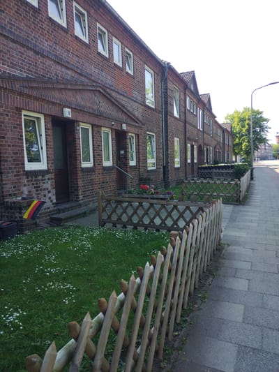 The image shows a row of brick apartment buildings with many windows. A sidewalk runs along the side of the buildings, and a small wooden fence separates the sidewalk from a grassy area with small white flowers. There is a German flag displayed on the grass near the entrance to one of the apartments. The scene appears to be in a residential area of Cuxhaven, Germany.