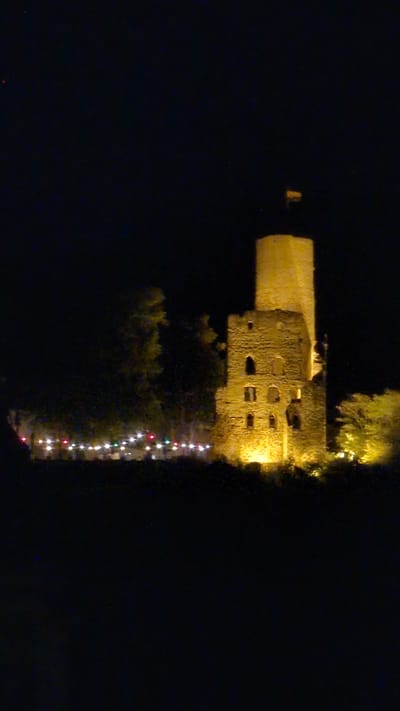 A medieval stone tower ruin, illuminated by artificial lights, stands tall against a black night sky. The tower has a cylindrical upper section and a more fragmented lower section with visible windows. In front of the tower, a line of colorful string lights is visible, suggesting an event or decoration. Trees and foliage surround the base of the tower, also partially lit. The scene is set at the Strahlenburg castle in Schriesheim, Germany, which is a landmark and ruin overlooking the town. There are no people or discernible activity depicted in the image.