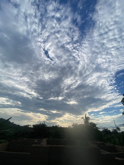 A wide-angle, upward-facing shot captures a dramatic sky over Ufu Esioni, Nigeria. The sky is filled with a complex pattern of clouds, ranging from dense, dark grey formations to wispy, white streaks that catch the sunlight. Patches of vibrant blue sky are visible between the clouds. In the lower portion of the image, a silhouette of lush green vegetation and palm trees forms a dark horizon. In the foreground, the rough, unfinished concrete structure of a building under construction is visible, with rebar protruding from some sections. The overall impression is one of an outdoor scene at dusk or dawn, with a construction site in the foreground and a dynamic sky above.