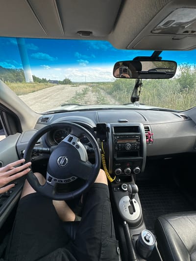 The image shows the interior of a car, likely a Nissan, with the driver's perspective. The view is oriented as if the car is parked and the photo is taken from the driver's seat looking forward and slightly to the right. The dashboard features a car stereo, climate controls, and a gear shift. A hand is visible on the steering wheel, suggesting the car is either parked or the driver is about to drive.

Outside the car, a bright blue sky with scattered clouds is visible through the windshield, indicating it is daytime. The landscape outside appears to be rural, with dry grass and a dirt road. The scene is set in Abrykosivka, Ukraine, as per the provided context.

Notable details include a rearview mirror reflecting a person holding a phone, a bright pink novelty item on the dashboard, and an open can of soda in the center console. The lighting suggests it's a sunny day. There are no discernible emotions conveyed in the image. The car's interior is functional, with visible wear and tear on some surfaces. The steering wheel has the Nissan logo in the center. There are no other people visible in the car besides the partial reflection in the rearview mirror.