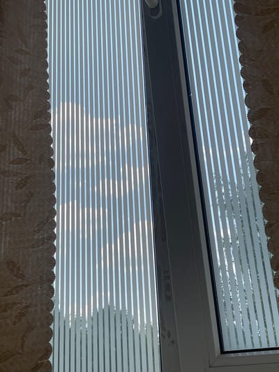 The image captures a view through a window in Tikhvin, Russia, adorned with vertical blinds. The blinds are partially open, allowing sunlight to stream through, casting striped shadows on what appears to be a wall or another surface not fully visible in the frame. The external environment is discernible as a bright sky with clouds and hints of trees and foliage, suggesting it is daytime and likely outdoors. The window frame itself is a neutral color, possibly white or gray, and shows some minor smudges or marks. On the left side of the image, a portion of a patterned curtain is visible, adding a textural element to the scene. The overall impression is that of a typical domestic or office window view.