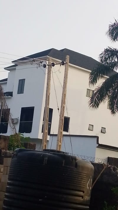 The image depicts a two-story building in Enugwu-Ukwu, Nigeria. The building is painted white with dark-framed windows and a dark roof. In the foreground, there is a large black plastic water tank. Two wooden utility poles stand between the building and the water tank, with electrical wires running across the top. A palm tree is visible on the right side of the image. A low fence with barbed wire runs along the base of the building. Construction materials, including concrete blocks, are scattered in the foreground. The sky is overcast.
