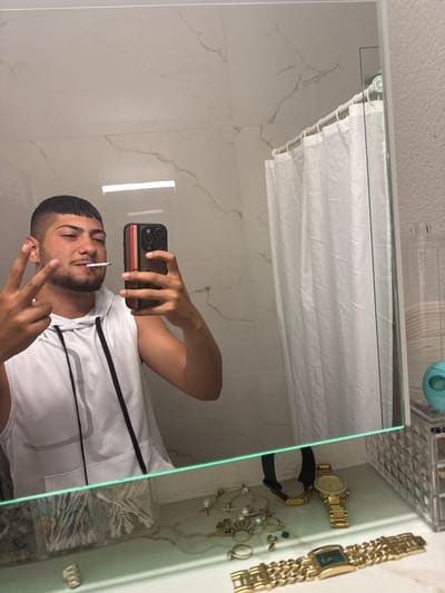 A man is taking a selfie in a bathroom mirror. He is wearing a white tank top with black drawstrings and holding a cigarette in his mouth. He is making a peace sign with his fingers. The background of the reflection includes a shower curtain, marble-patterned walls, and a shelf with jewelry and cotton swabs. There is no evidence to suggest that the location is Lausen, Switzerland.