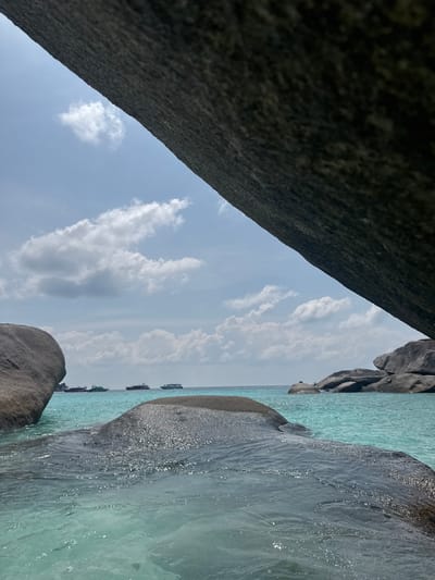 The image captures a view from what appears to be a rocky outcrop in Lam Kaen, Thailand. The foreground is dominated by large, dark gray rocks forming a natural frame. Between the rocks is a view of the turquoise sea and a partly cloudy sky. Several boats can be seen on the horizon. Sunlight glints off the water, creating a shimmering effect.