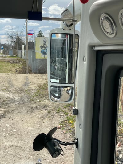 The image provides an interior view from a vehicle, likely a bus or large van, looking out through the front passenger-side window. The perspective is slightly angled, capturing both the vehicle's internal structure and the exterior environment.

**Subjects:** In the foreground, the vehicle's white overhead console and ceiling are visible, featuring two circular air vents. To the right, a black fan-like device is suspended from the ceiling. A side mirror reflects parts of the vehicle's interior, showing items covered in protective plastic, along with a glimpse of the ground behind. Outside the window, the scene includes an unpaved dirt road, patches of green grass, and some yellow vegetation. Utility poles with wires and bare trees, indicating late winter or early spring, line the road. In the midground, a large yellow billboard with a graphic of a stylized globe and a building is prominently displayed. Adjacent to the billboard, a flag flies on a flagpole.

**Activity or Event:** The vehicle appears to be stationary or moving slowly along the unpaved road.

**Notable Details:** The time of day is bright and sunny, with a clear blue sky dotted with scattered white clouds. A significant location cue is the flag, which features horizontal white, blue, and red stripes with a distinctive wavy blue line and Cyrillic text. The text on the flag reads "ДОБРО ПОЖАЛОВАТЬ В РК", translating to "Welcome to RK". Given the location context of Bol'shoe Teshkovo, Russia, "RK" likely refers to the Republic of Crimea.
