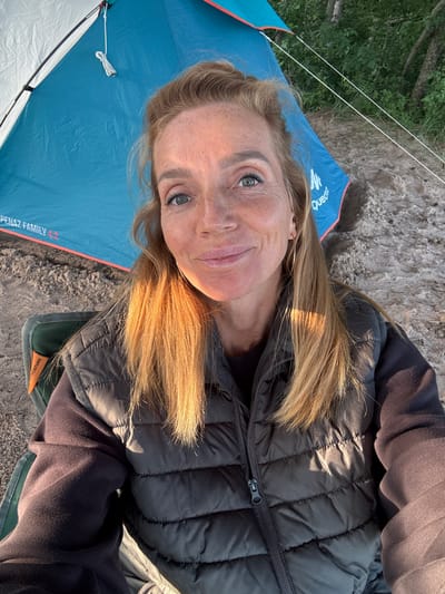 In Shepelevo, Russia, a woman is seated in a folding camping chair next to a blue and white tent. She is wearing a dark gray puffer vest over a dark purple hoodie. Her light brown hair is shoulder-length, and she is looking at the camera with a slight smile. The tent is set up on a sandy surface, with trees visible in the background. The sun is casting warm light on the scene, highlighting the woman's face and hair.