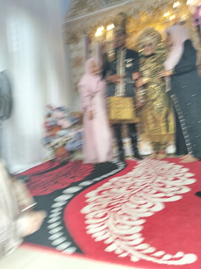 A blurry, motion-filled image depicts a wedding ceremony in a decorated venue, likely in Cottrueng, Indonesia. In the center, a man in a dark, sparkly suit stands next to a woman wearing a shimmering gold dress and a large blonde wig. To their right, another woman is dressed in a dark outfit and a light pink headscarf. To the left, a person in a light pink garment, possibly a bridesmaid or guest, stands slightly apart. The individuals appear to be interacting, but their precise actions are obscured by motion blur. The background is dimly lit and features decorative elements, including what appears to be a draped canopy and lights. In the foreground, a red carpet with a white floral or paisley pattern is visible, suggesting a formal setting. To the far left, a portion of a fan and some floral arrangements are also discernible. The overall impression is one of a vibrant, albeit blurry, celebratory event.