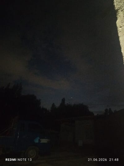 A nighttime exterior scene captured in Асан-Чек, Kyrgyzstan, features a dark, cloudy sky with a single bright point, possibly a star or planet, visible in the upper center. Silhouetted trees form a dark line across the midground. In the foreground, a dark-colored pickup truck is parked on the left, with its wheel and part of the bed visible. To the right of the truck is a low-rise building, possibly a shed or garage, with a faint light or window glowing from within. The overall image is dark and grainy, indicative of a low-light photograph. The bottom left corner displays "REDMI NOTE 13" and the bottom right corner shows the date "21.06.2026" and time "21:48". A textured, light-colored surface, possibly a wall or building facade, is visible along the far right edge of the frame.