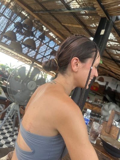 The image presents a profile view of a person, likely female, captured from the right side, looking upwards and slightly to the right. She has dark hair, styled with two thin braids framing her face, one adorned with a small silver clover-shaped charm. Her hair is tied in a low bun at the back. Her mouth is slightly open, revealing teeth, indicating a smile or laughter. A thin gold chain necklace is visible around her neck, and a faint tattoo is on her upper back. She is wearing a light gray, ribbed top with cut-out details on the side.
The setting appears to be an open-air establishment, possibly a cafe or restaurant, characterized by a rustic, woven, or thatched ceiling structure that allows light to filter through. Several items are visible in the background: a large standing fan (brand "Senko" visible), a tabletop with a glass containing ice and a dark drink, and a plastic "AQUAFINA" water bottle. The floor features a distinct black and white geometric tiled pattern. In the blurred background, a red object with a yellow star is visible, consistent with the flag of Vietnam, confirming the location context of Hà Giang 1 Ward, Vietnam. The scene conveys a relaxed and pleasant moment, likely during the day in a warm environment.