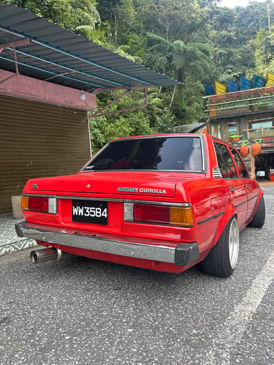 A rear, slightly low-angle, full shot captures a bright red, vintage Toyota Corolla sedan parked on a paved road with a white lane marker visible on the right. The car is positioned at an angle, facing away from the viewer and slightly to the left. Its rear bumper is chrome with black corner guards, and a visible exhaust pipe extends from the left side. The license plate, "WW3584", is prominently displayed in white on a black background. The car has wide tires and chrome rims.
Behind the car, a building with a corrugated metal roof and pink-colored walls is visible. To the right of this building, another structure with a colorful facade, featuring yellow and blue triangular accents and large decorative strawberries, is present. This structure appears to be a shop or cafe.
The background is dominated by lush, green vegetation and trees, indicative of a hilly or mountainous area, suggesting a location in or near the Cameron Highlands, possibly in Tanah Rata, Malaysia. The sky is overcast, and the lighting is diffused. No people are visible in the frame.
