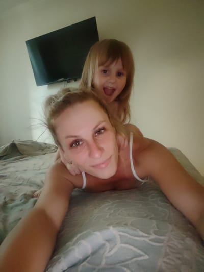 A selfie is taken from a low angle, showing a woman lying on her front on a bed with a young child on her back, also facing the camera. The woman has blonde hair pulled back in a bun and is wearing a white tank top. Her eyes are brown, and she is smiling. The child has blonde hair and is also smiling with an open mouth. Behind them, a black flat-screen television is mounted on a light-colored wall. The bed is covered with a light blue patterned sheet. There is no identifiable information to place this image in Mezhdurechensk, Russia.