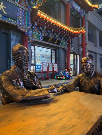 The image shows two bronze statues of men, one sitting and one standing, appearing to shake hands across a wooden surface. Behind the statues is a building in Linghe, China, decorated in a traditional Chinese architectural style with red pillars and ornate carvings, illuminated by string lights. There is a shop visible with window displays and various objects, including what appears to be a child's ride. The backdrop includes modern buildings. The image has a vertical orientation.