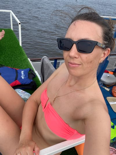 A woman in a pink bikini and sunglasses is sitting on a boat on the water in Golyanovo, Russia. She is holding a bottle of water and a life vest is next to her. The water is dark and choppy.