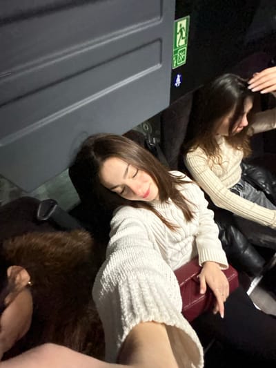 Two women are reclining on a dark surface, possibly seats. Both wear light-colored, textured sweaters. One woman lies atop a burgundy purse. The first woman has long brown hair and closed eyes. A dark brown, possibly fur, item is next to her. A grey panel is in the bottom left with emergency exit signs visible. A second woman is further back, with dark leather-like pants visible. The image has a slightly tilted perspective.